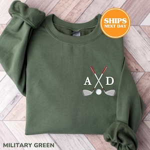 Puede incluir: Una sudadera de cuello redondo color verde militar con un diseño bordado en el pecho izquierdo. El diseño incluye dos palos de golf cruzados con empuñaduras rojas, una pelota de golf blanca y las letras mayúsculas blancas A y D a los lados.