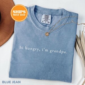 May include: A blue jean colored crew neck t-shirt with the white text "hi hungry, i'm grandpa." printed on the chest. The shirt features a Comfort Colors brand label at the inner neckline and is paired with a delicate gold-toned chain necklace.