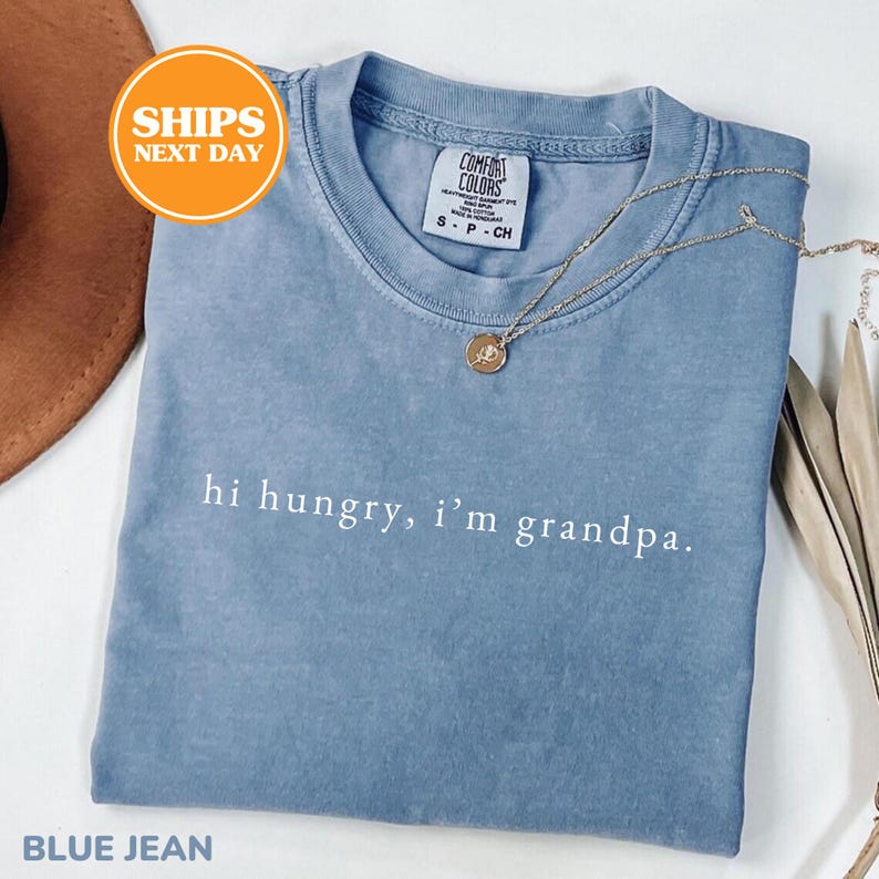 May include: A blue jean colored crew neck t-shirt with the white text "hi hungry, i'm grandpa." printed on the chest. The shirt features a Comfort Colors brand label at the inner neckline and is paired with a delicate gold-toned chain necklace.