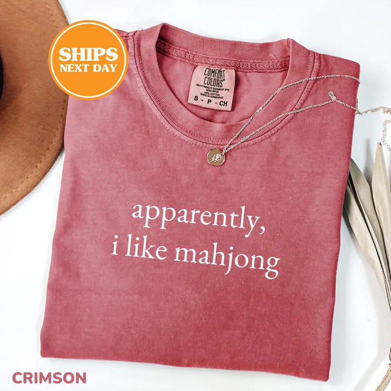 May include: Crimson-colored t-shirt with the text "apparently, i like mahjong" in white. A silver necklace with a pendant is draped across the shirt. The shirt is folded and displayed on a white surface.