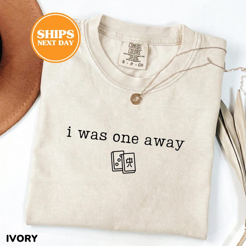 May include: An ivory-colored crew neck t-shirt featuring the black text "i was one away" printed above a graphic of two mahjong tiles. The shirt has a Comfort Colors brand label at the inner neckline and is paired with a thin gold-toned necklace.