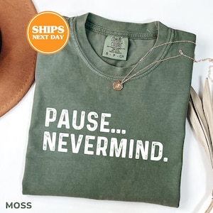 May include: A moss green short-sleeved t-shirt with a crew neckline and a distressed white graphic print that reads PAUSE... NEVERMIND. The shirt features a Comfort Colors brand label at the inner neck and is paired with a delicate gold-toned necklace.