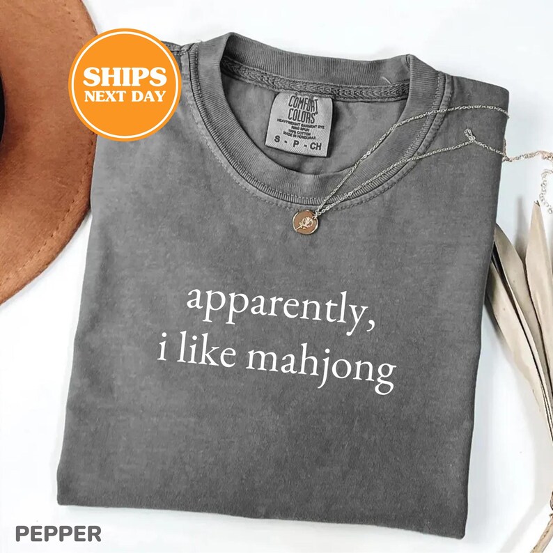 May include: A gray t-shirt with the text "apparently, i like mahjong" in white. The shirt has a crew neck and short sleeves. A silver necklace with a round pendant rests on the shirt. The shirt is folded on a white surface.