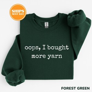 A green sweatshirt with white text that says "oops, I bought more yarn" and an orange circular sticker that says "SHIPS NEXT DAY".