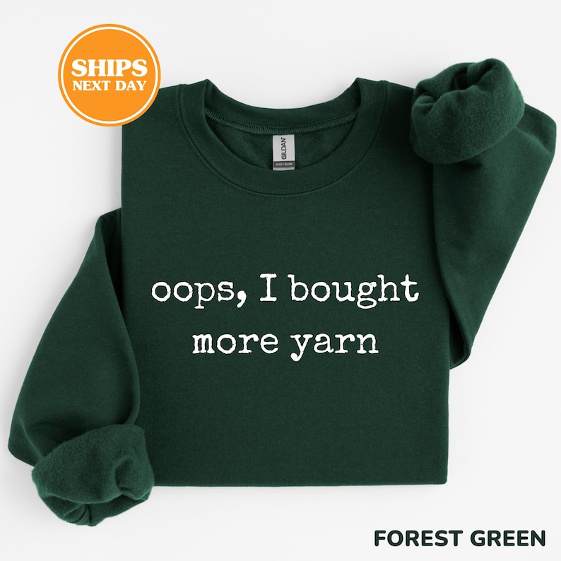 A green sweatshirt with white text that says "oops, I bought more yarn" and an orange circular sticker that says "SHIPS NEXT DAY".