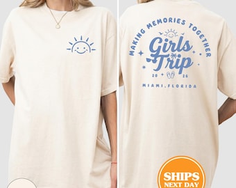 Könnte beinhalten: Elfenbeinfarbenes T-Shirt mit blauem Sonnensymbol auf der Vorderseite. Auf der Rückseite steht in Blau "MAKING MEMORIES TOGETHER Girls Trip 20 26 MIAMI, FLORIDA". Das Shirt hat kurze Ärmel und eine lockere Passform.