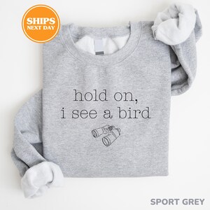 A gray sweatshirt with the text "hold on, I see a bird" printed on it, along with a small illustration of binoculars.