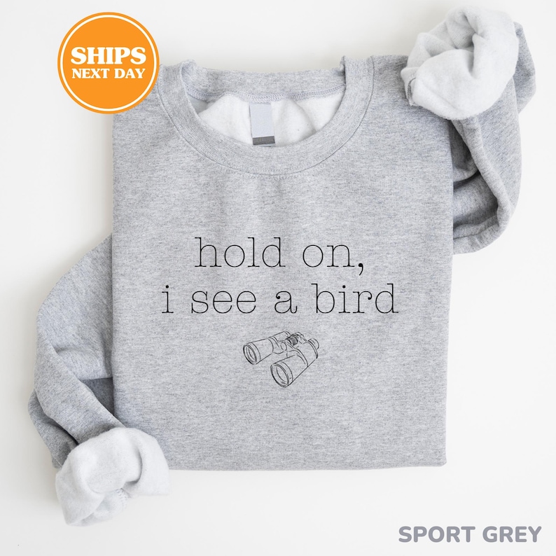 A gray sweatshirt with the text "hold on, I see a bird" printed on it, along with a small illustration of binoculars.