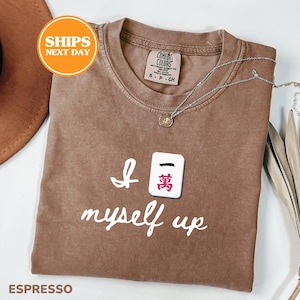Pu&ograve; includere: T-shirt color espresso con la frase "I myself up" e una grafica a piastrella Mahjong. La maglietta ha un girocollo e maniche corte. Una collana d'argento con un ciondolo &egrave; drappeggiata sulla maglietta.