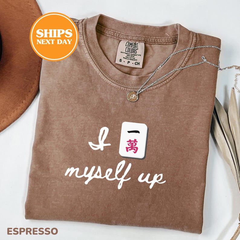 Pu&ograve; includere: T-shirt color espresso con la frase "I myself up" e una grafica a piastrella Mahjong. La maglietta ha un girocollo e maniche corte. Una collana d'argento con un ciondolo &egrave; drappeggiata sulla maglietta.