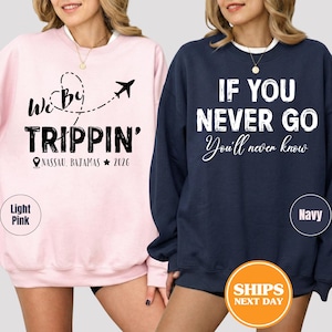 May include: Two sweatshirts, one light pink and one navy blue. The light pink sweatshirt has the text "We By TRIPPIN'" with a plane graphic and "NASSAU, BAJAMAS 2026". The navy blue sweatshirt says "IF YOU NEVER GO You'll never know".