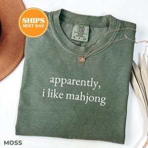 May include: A moss green t-shirt with the text "apparently, i like mahjong" in white. The shirt has a crew neck and short sleeves. A gold necklace with a pendant is draped on the shirt. The shirt is labeled "Comfort Colors".