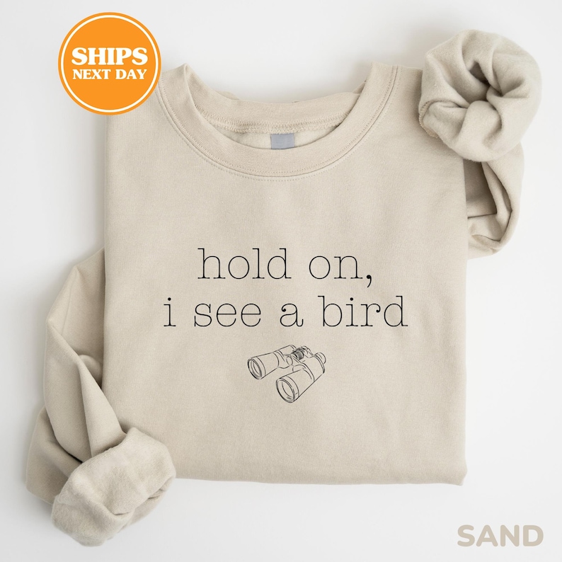 A beige t-shirt with the text "hold on, I see a bird" printed on it, along with a small illustration of binoculars.
