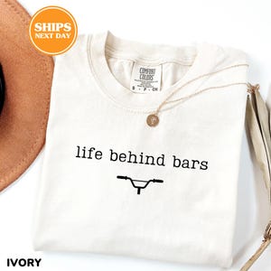 a white t-shirt with the text "life behind bars" printed on it, along with a bicycle graphic. The t-shirt is laid out on a white surface, and there is a brown hat and a necklace visible in the background.
