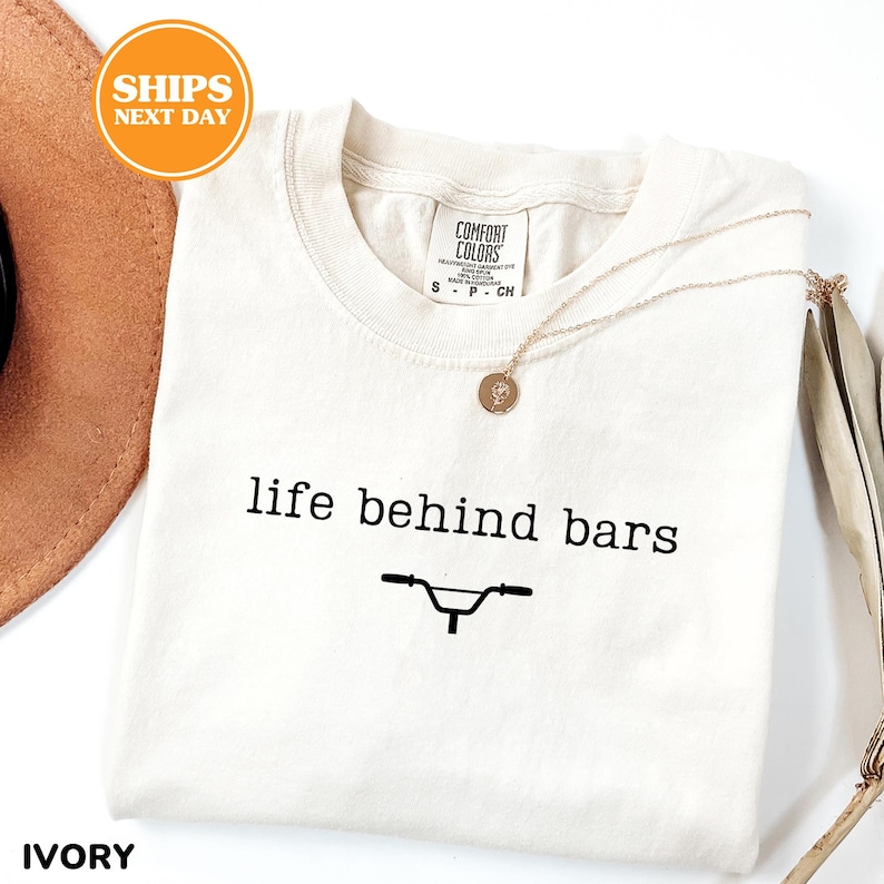 a white t-shirt with the text "life behind bars" printed on it, along with a bicycle graphic. The t-shirt is laid out on a white surface, and there is a brown hat and a necklace visible in the background.