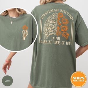 May include: Moss green t-shirt with a skeleton hand holding orange flowers on the front. The back features a rib cage design with orange flowers and the text "FIND SOMEONE WHO GROWS FLOWERS IN THE DARKEST PARTS OF YOU."