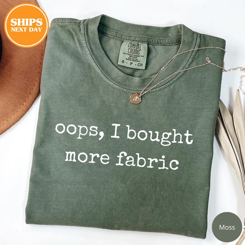 A green t-shirt with the text "oops, I bought more fabric" printed on it, along with a small charm or pendant. The t-shirt is laid out on a white surface, and there is a brown hat and some leaves in the background.
