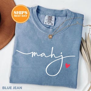 May include: A blue jean colored t-shirt with the word "mahj" in white script and a small red heart. A gold necklace with a pendant is draped across the shirt. The shirt is folded and has a "Comfort Colors" tag.