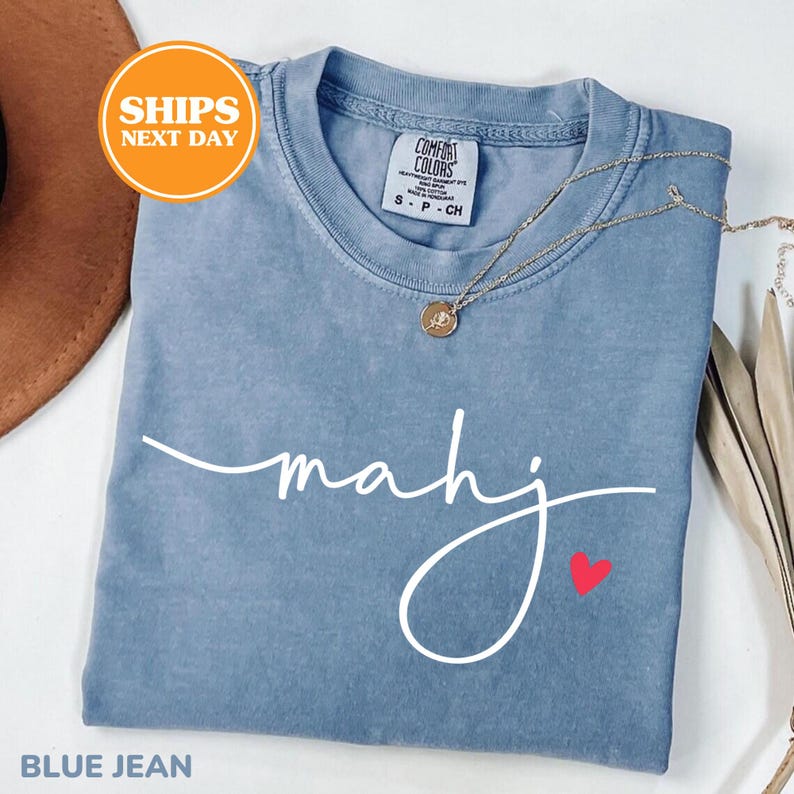 May include: A blue jean colored t-shirt with the word "mahj" in white script and a small red heart. A gold necklace with a pendant is draped across the shirt. The shirt is folded and has a "Comfort Colors" tag.