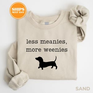 A beige t-shirt with the text "less meanies, more weenies" printed on it, along with a silhouette of a dog.