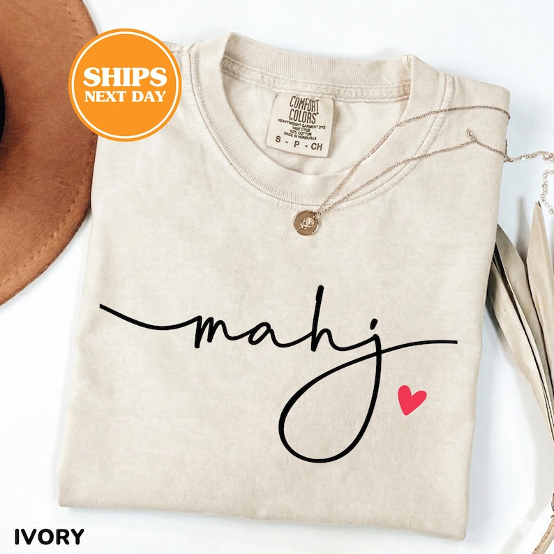 May include: Ivory-colored t-shirt with the word "mahj" in black script and a small red heart. The shirt is a Comfort Colors brand and has a crew neck. A gold necklace is draped across the shirt.