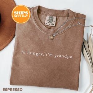 May include: A brown short-sleeved t-shirt with a crew neckline and the white text "hi hungry, i'm grandpa." printed on the chest. The shirt features a Comfort Colors brand label inside the collar and a thin silver-toned chain necklace.