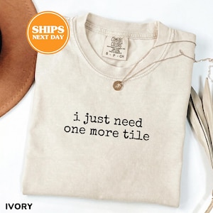 May include: Ivory-colored t-shirt with the text "i just need one more tile" in black. The shirt has a crew neck and short sleeves. A gold necklace with a pendant is draped across the shirt. The shirt is labeled "Comfort Colors".