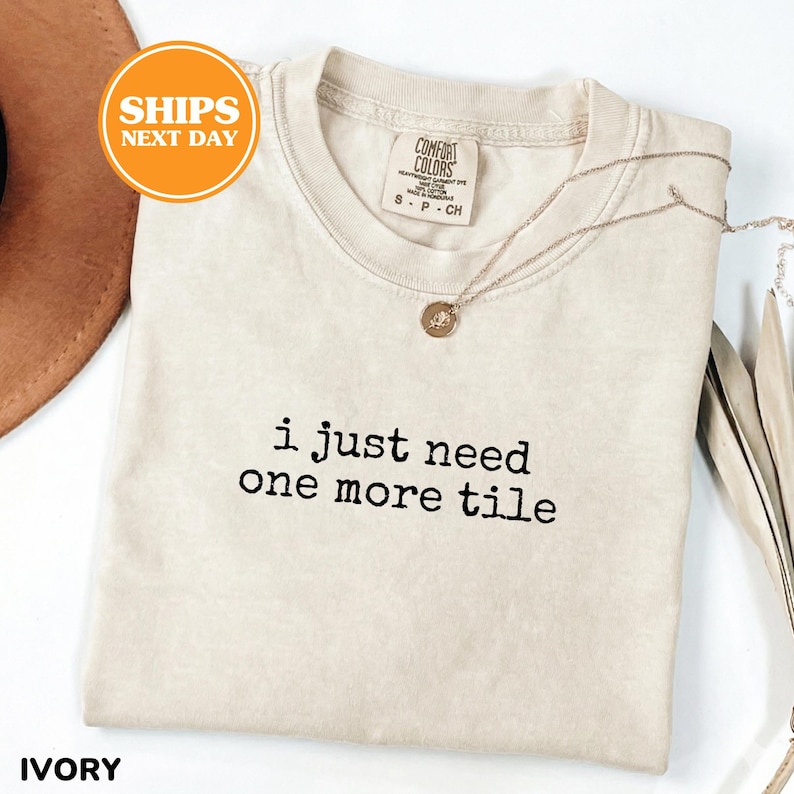 May include: Ivory-colored t-shirt with the text "i just need one more tile" in black. The shirt has a crew neck and short sleeves. A gold necklace with a pendant is draped across the shirt. The shirt is labeled "Comfort Colors".