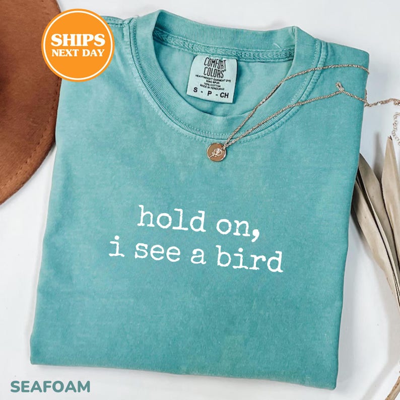 A turquoise t-shirt with the text "hold on, I see a bird" printed on it, along with a necklace and a brown hat.