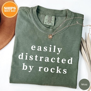 May include: Moss green t-shirt with the text "easily distracted by rocks" in white. The shirt has a crew neck and short sleeves. A gold necklace with a pendant is draped on the shirt.