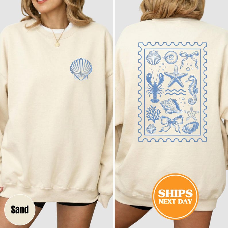 May include: A cream-colored crewneck sweatshirt featuring a small blue shell graphic on the front left chest. The back displays a large rectangular stamp-style design containing blue illustrations of a lobster, starfish, seahorse, and shells.