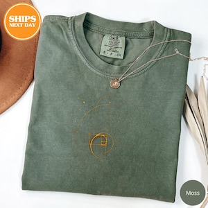 May include: A moss green Comfort Colors t-shirt with a gold spiral design. The shirt has a crew neck and short sleeves. A silver necklace with a small pendant rests on the shirt. The shirt is folded and has a small tag.