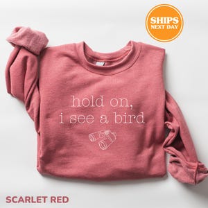 A red sweater with the text "hold on, I see a bird" printed on it, along with a pair of binoculars. The sweater is folded and placed on a white background, with a "SHIPS NEXT DAY" sticker visible in the top right corner.