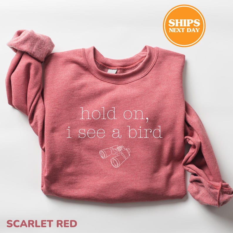 A red sweater with the text "hold on, I see a bird" printed on it, along with a pair of binoculars. The sweater is folded and placed on a white background, with a "SHIPS NEXT DAY" sticker visible in the top right corner.
