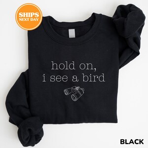 A black sweatshirt with the text "hold on, I see a bird" printed on it, along with a small illustration of binoculars.