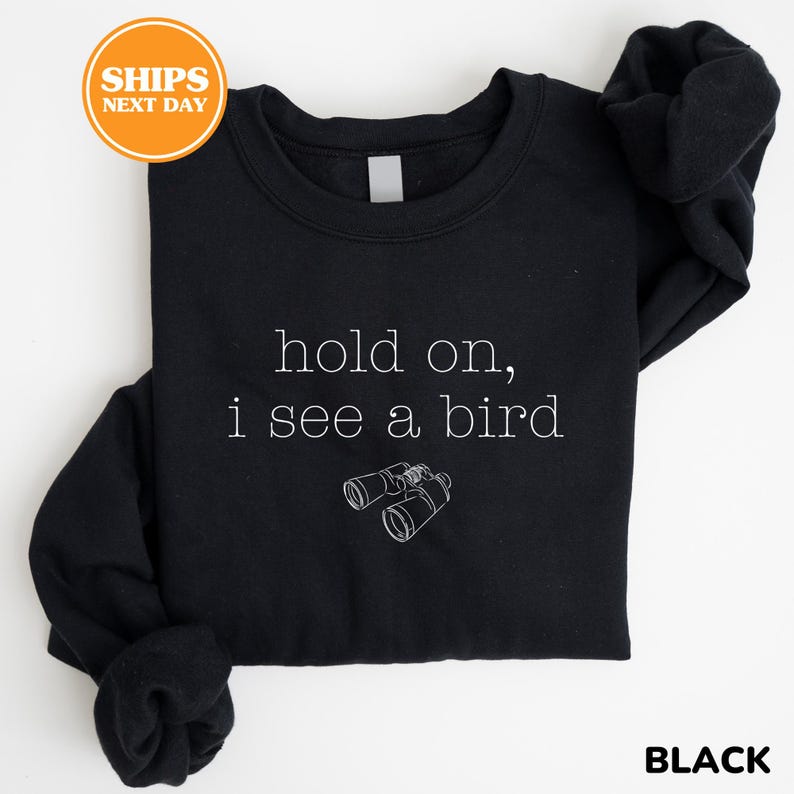 A black sweatshirt with the text "hold on, I see a bird" printed on it, along with a small illustration of binoculars.