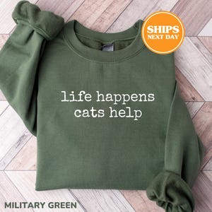 May include: A military green crewneck sweatshirt featuring the white text "life happens cats help" printed in a typewriter-style font across the chest. The garment has long sleeves with ribbed cuffs and a ribbed neckline.