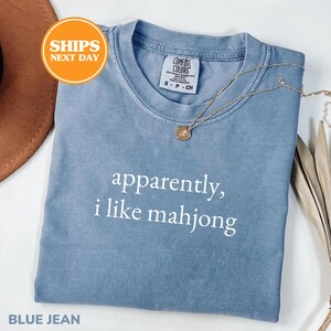 May include: A blue jean colored t-shirt with the text "apparently, i like mahjong" in white. The shirt has a crew neck and short sleeves. A gold necklace with a pendant is draped on the shirt.