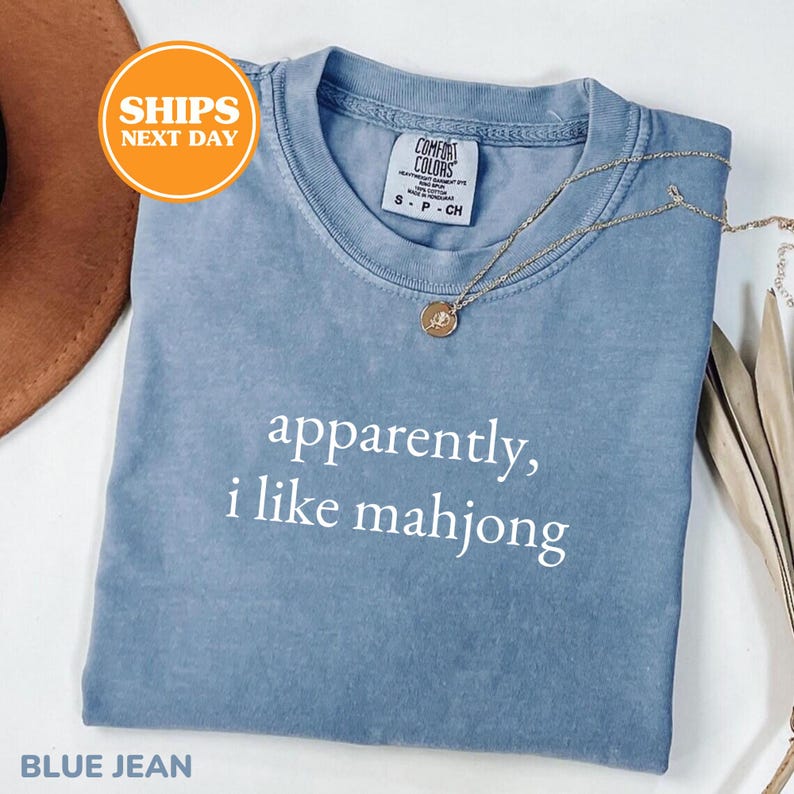 May include: A blue jean colored t-shirt with the text "apparently, i like mahjong" in white. The shirt has a crew neck and short sleeves. A gold necklace with a pendant is draped on the shirt.