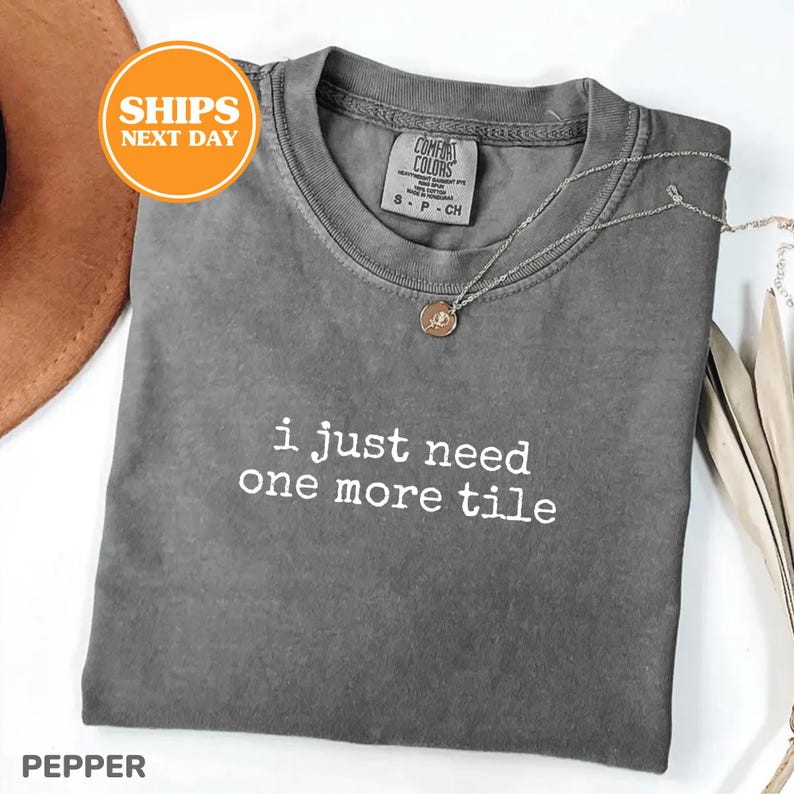 May include: A gray Comfort Colors t-shirt with the text "i just need one more tile" in white. A silver necklace with a pendant rests on the shirt. The shirt is folded and displayed with a brown hat.