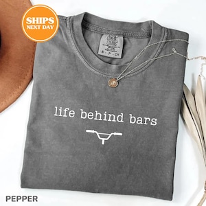 a gray t-shirt with the text "life behind bars" printed on it, along with a graphic of a bicycle. The t-shirt is laid out on a white surface, and there is a brown hat and some leaves in the background.