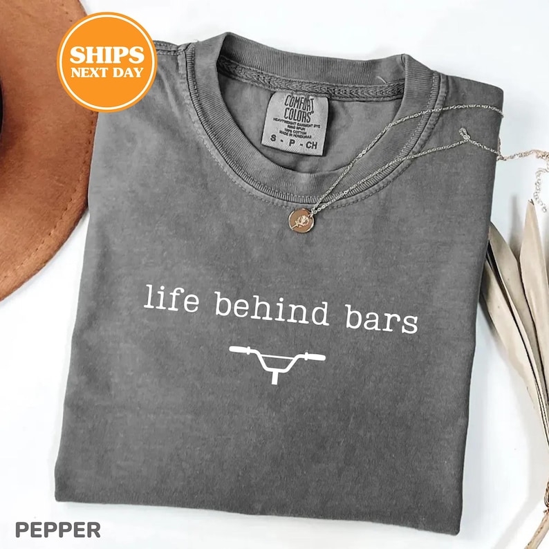 a gray t-shirt with the text "life behind bars" printed on it, along with a graphic of a bicycle. The t-shirt is laid out on a white surface, and there is a brown hat and some leaves in the background.