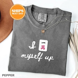 Pu&ograve; includere: Una maglietta grigia con la scritta "I myself up" e una grafica a piastrella Mahjong. Una collana d'argento con un ciondolo &egrave; drappeggiata sulla maglietta. La maglietta &egrave; piegata e presentata su una superficie bianca.
