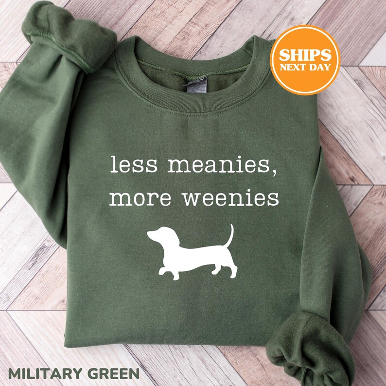 A green sweatshirt with the text "less meanies, more weenies" printed on it, along with a white silhouette of a dog.