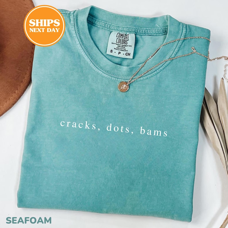 A turquoise t-shirt with the text "cracks, dots, bams" printed on it, along with a necklace and a brown hat.