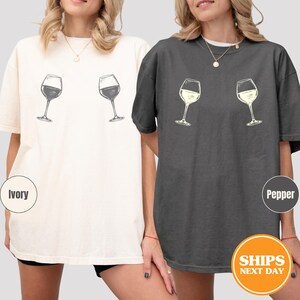 May include: Two t-shirts, one ivory and one pepper, each featuring a sketch of two wine glasses. The ivory shirt has dark wine glasses, while the pepper shirt has light wine glasses. The shirts are casual and comfortable.