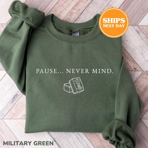 May include: A military green crewneck sweatshirt featuring the white text PAUSE... NEVER MIND. centered on the chest. Below the text is a simple white line art illustration of two mahjong tiles, one of which displays a character symbol.
