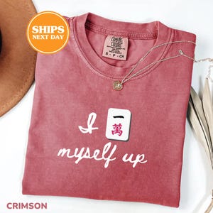 Pu&ograve; includere: T-shirt color cremisi con la scritta "I myself up" e una grafica a piastrella Mahjong. La maglietta ha uno scollo tondo e una piccola etichetta sul colletto. Una collana d'argento con un ciondolo &egrave; drappeggiata sulla maglietta.