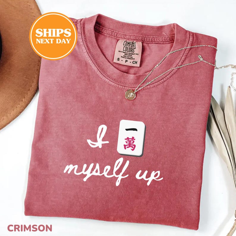 Pu&ograve; includere: T-shirt color cremisi con la scritta "I myself up" e una grafica a piastrella Mahjong. La maglietta ha uno scollo tondo e una piccola etichetta sul colletto. Una collana d'argento con un ciondolo &egrave; drappeggiata sulla maglietta.