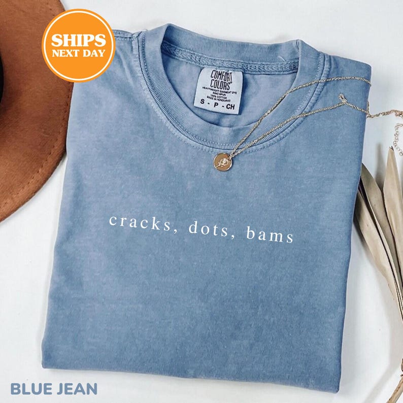 A blue t-shirt with the text "cracks, dots, bams" printed on it, along with a necklace and a brown hat.